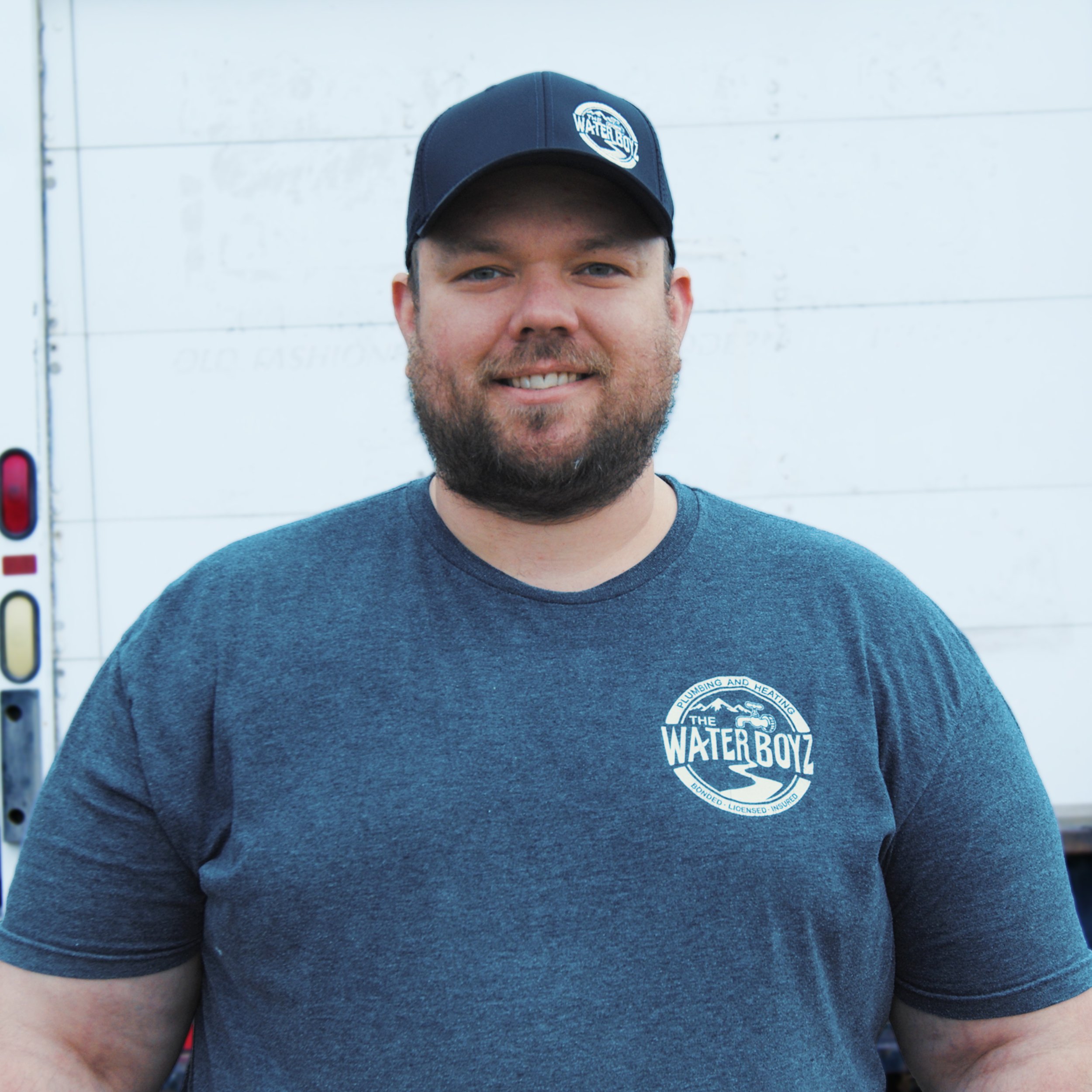 Close-up of a man wearing a gray T-shirt with a circular logo and a black baseball cap with a logo, standing against a white wall background.
