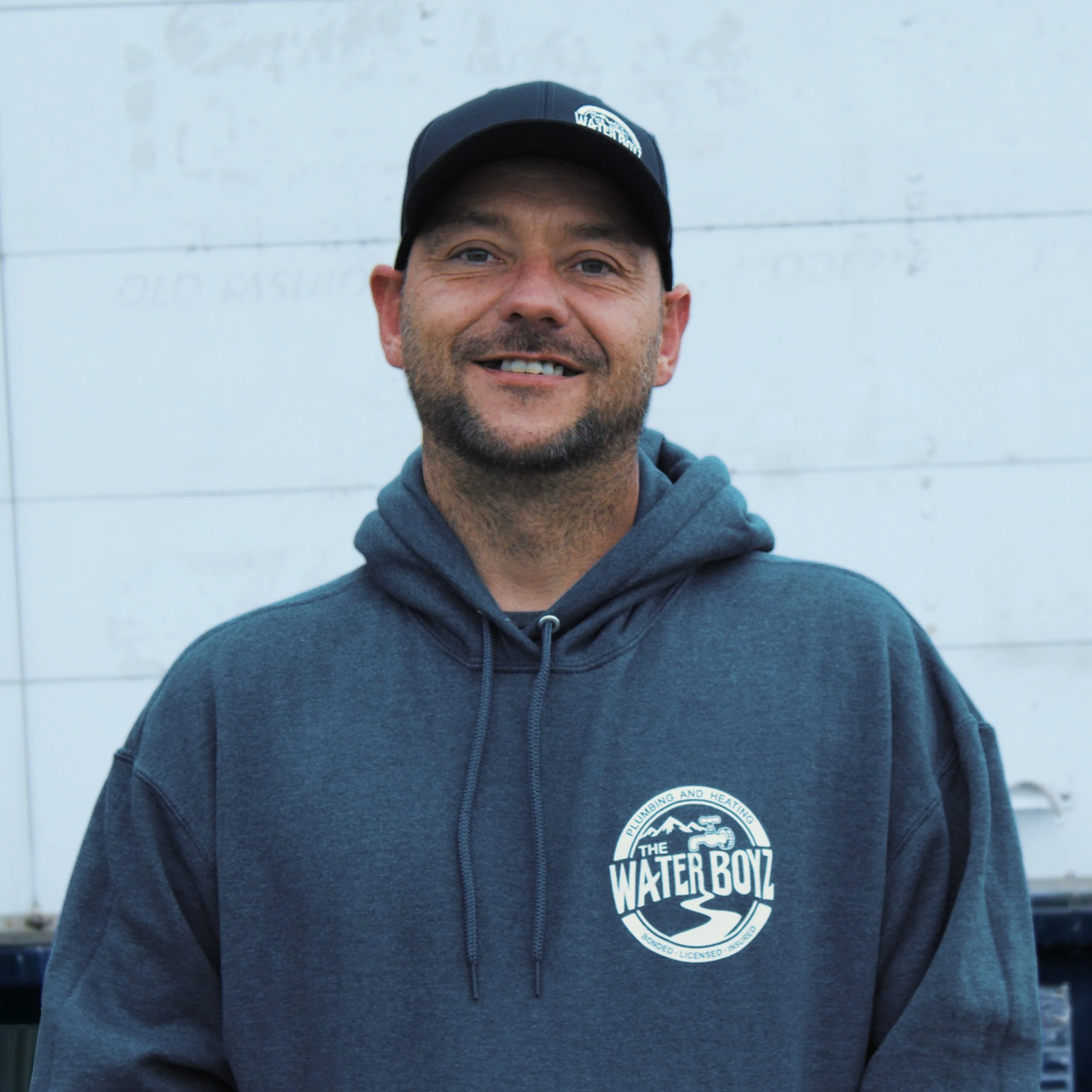 Close-up of a smiling man wearing a dark blue hoodie and a black cap with a logo. The logo on the hoodie and cap reads 'The Water Boyz' with an image of water waves and a water drop. The background is a white wall with faint horizontal lines.