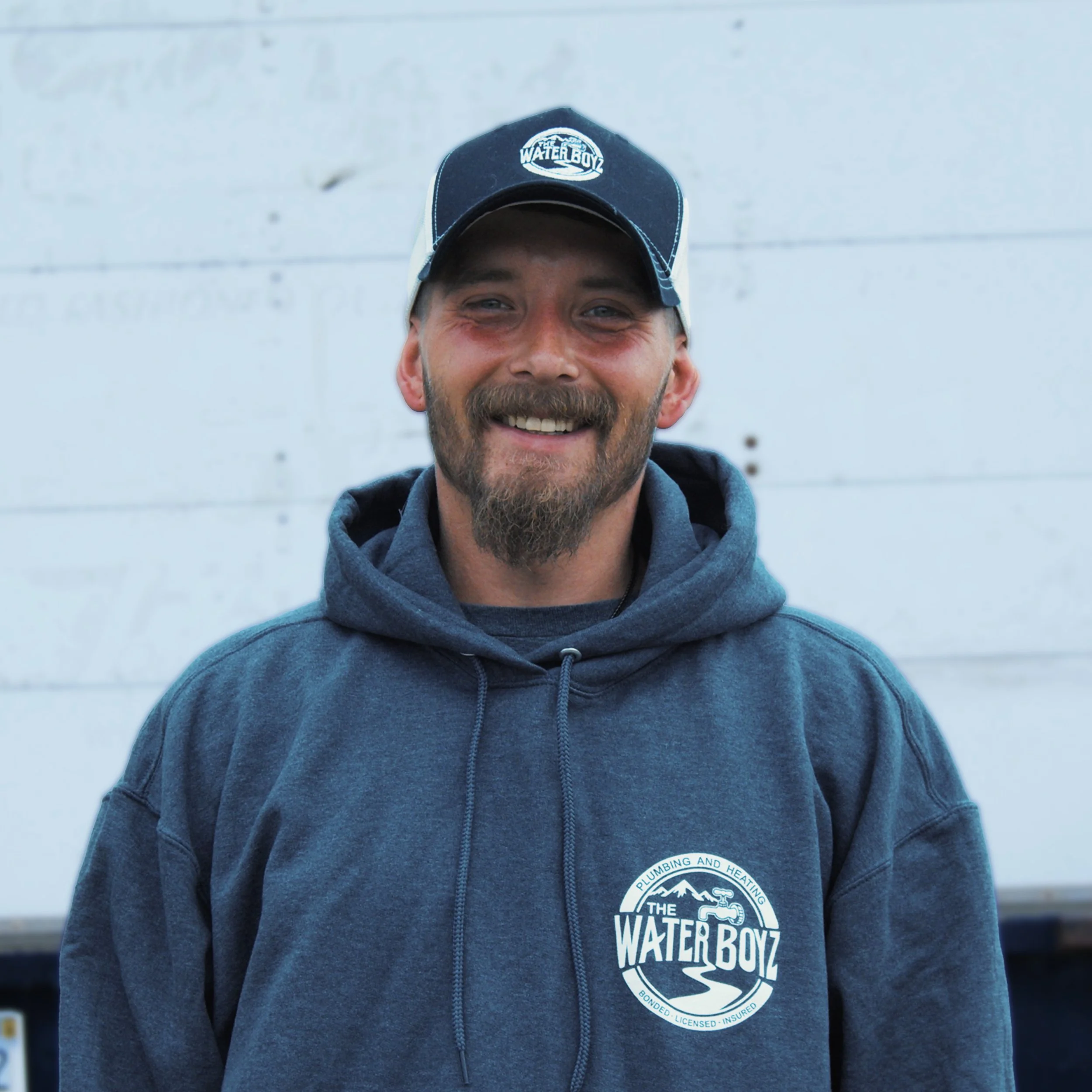 A man smiling wearing a black and white baseball cap and a gray hoodie with a logo reading 'The Water Boyz - Bonded, Licensed, Insured' against a light background.