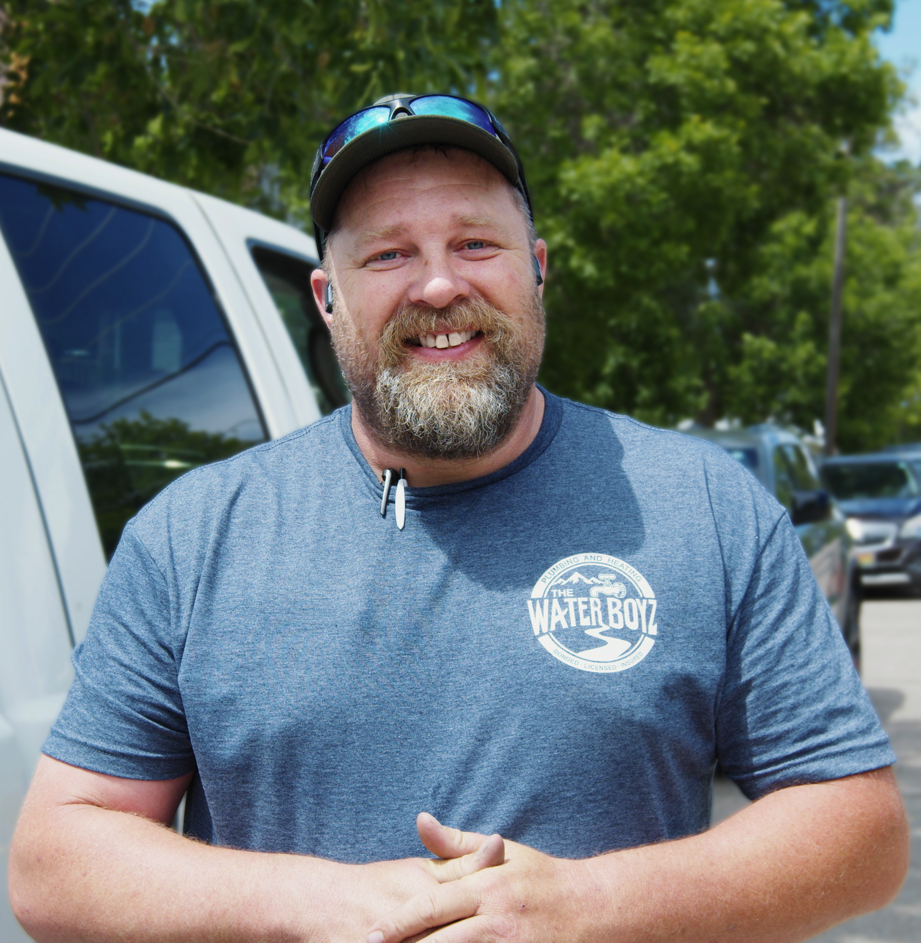 Smiling man with sunglasses on cap, beard, wearing a blue Water Boyz t-shirt, standing outdoors in front of a white vehicle and green trees.