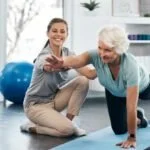 A young woman and an elderly woman smiling and giving each other a high-five during a fitness or physical therapy session.