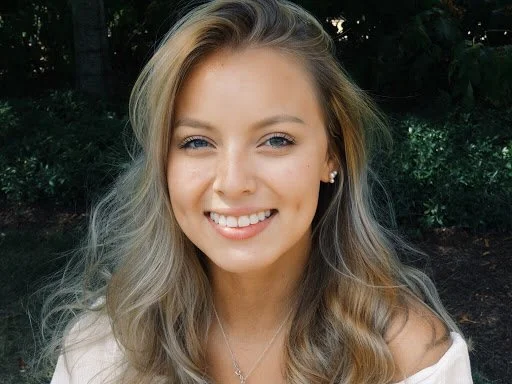 A young woman with long blonde hair, smiling outdoors with trees in the background.