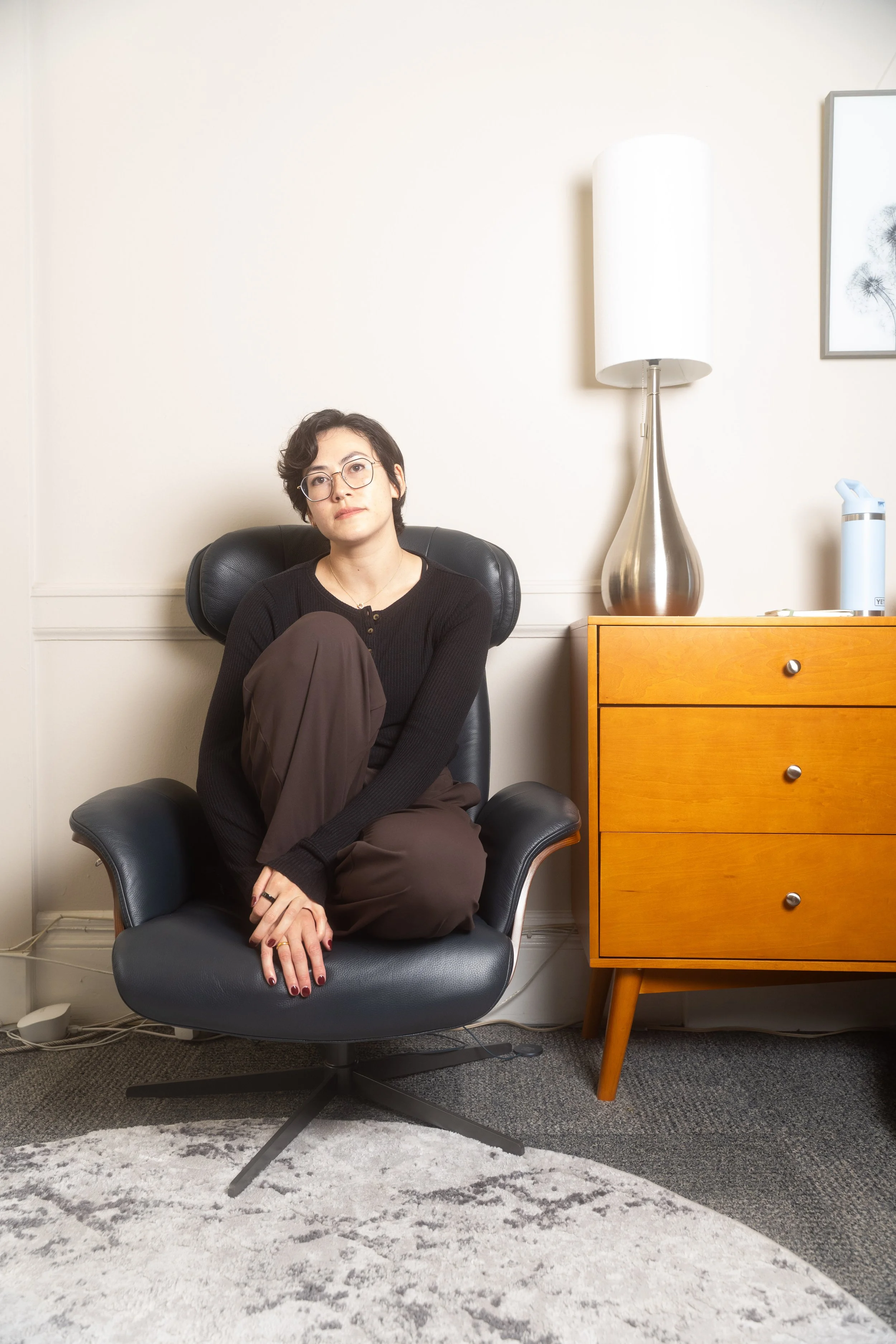 A woman with short dark hair, glasses, wearing a black cardigan and brown pants, sitting cross-legged in a black office chair in a room with beige walls, next to a wooden dresser with three drawers, a silver lamp, and a framed black-and-white artwork of dandelions on the wall.