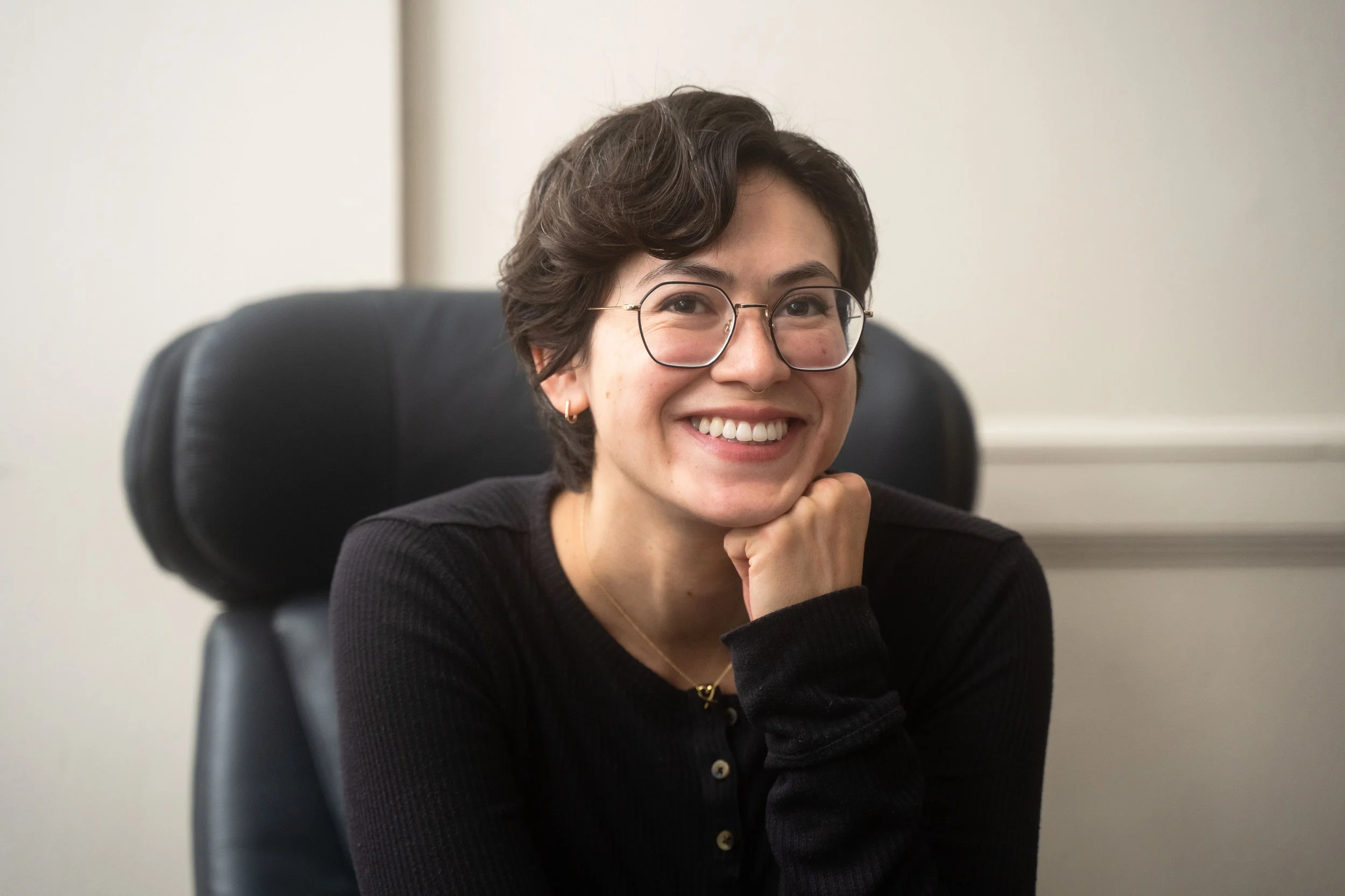 A woman with short dark hair, glasses, and a black top smiling while sitting in a black office chair.