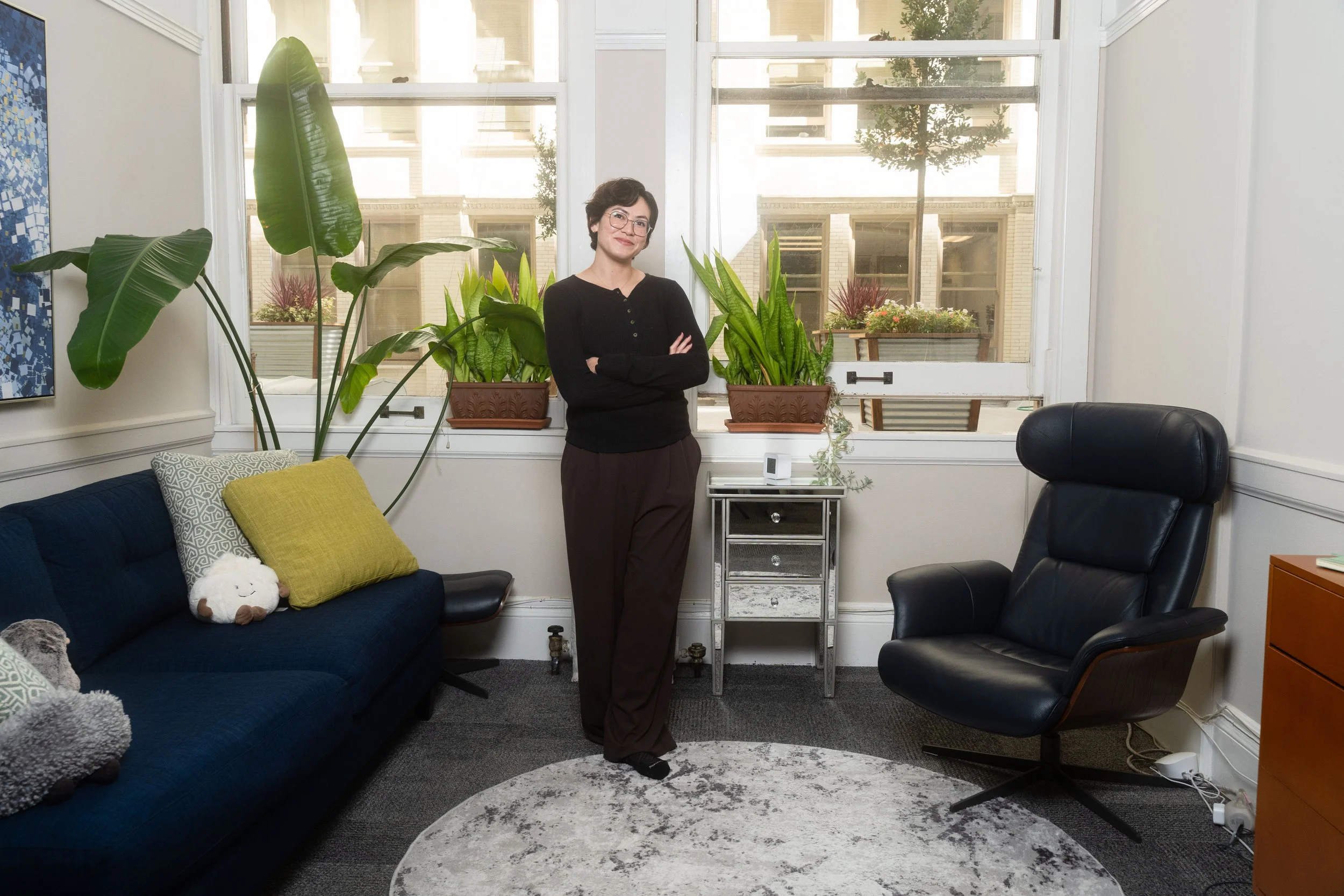 Interior of an office with a woman standing near a large window, surrounded by indoor plants, a blue sofa with cushions, and a black leather office chair.