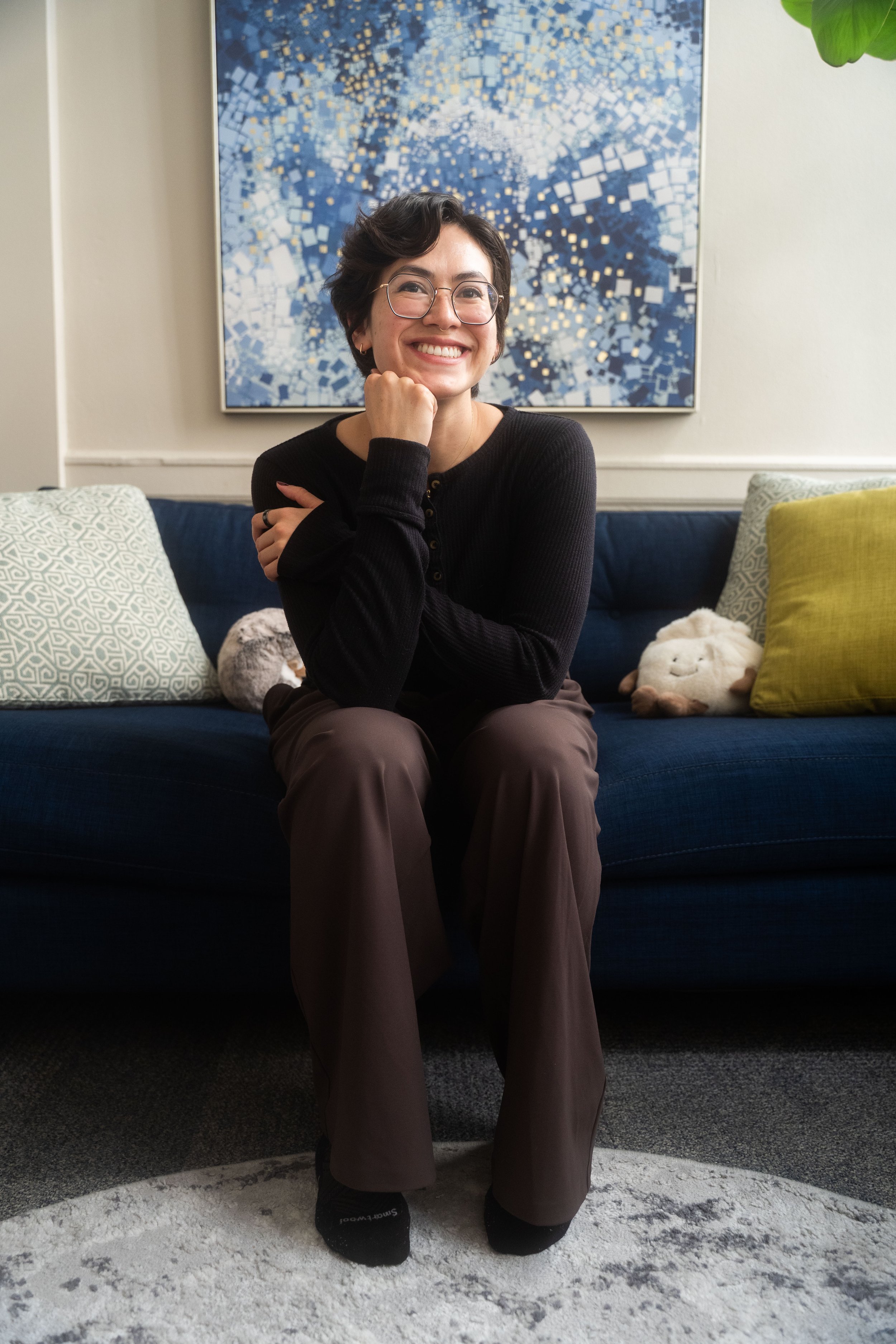 A woman with short dark hair, glasses, and a black long sleeve shirt sitting on a dark blue couch in a living room. She is smiling, resting her chin on her right hand, and wearing brown pants. Behind her is a large abstract painting with blue, white, and gold geometric shapes, and decorative pillows including a white pillow with a geometric pattern, a beige furry pillow, and a green pillow. There is also a plush animal toy and a plant with large green leaves visible to the right.