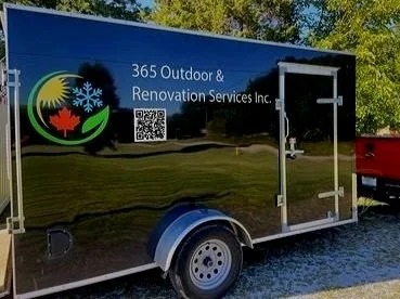 A black enclosed trailer with a logo and text for 365 Outdoor & Renovation Services Inc., parked outdoors on grass and gravel, with trees in the background.