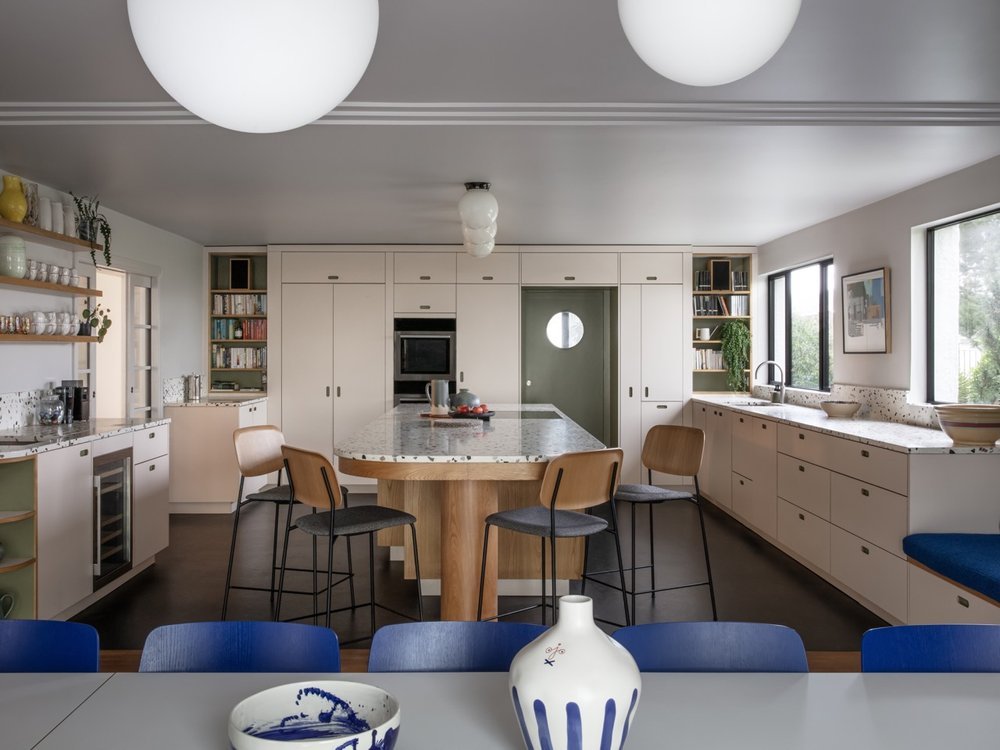 Modern kitchen with white cabinetry, a large central island, bar stools, built-in oven, open shelves with dishes, and large windows
