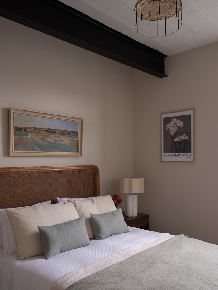A bedroom with beige walls, a wooden headboard, a white bed with light-colored bedding and pillows, a nightstand with a beige lamp, and framed artwork on the walls.