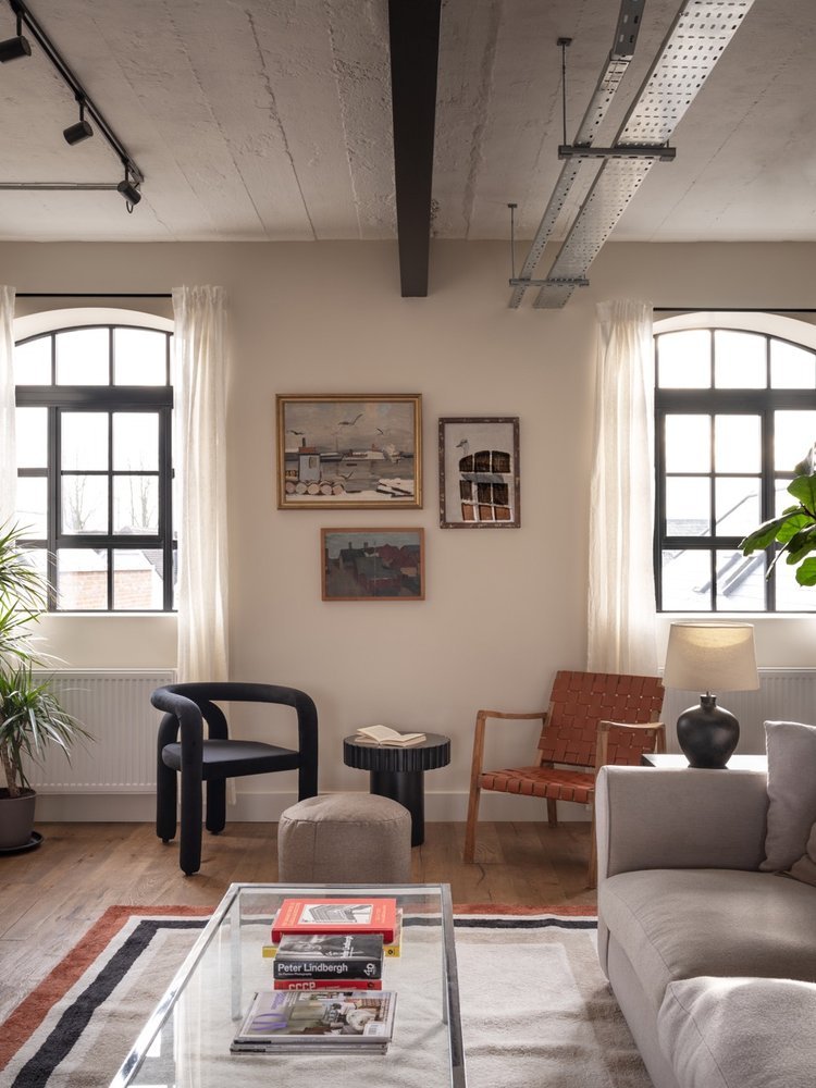 Living room with two large arched windows with curtains, a beige sofa, a wooden armchair with a red woven pattern, a black round side table with a book, a gray ottoman, a small modern black chair, a glass coffee table with books, framed artwork on th