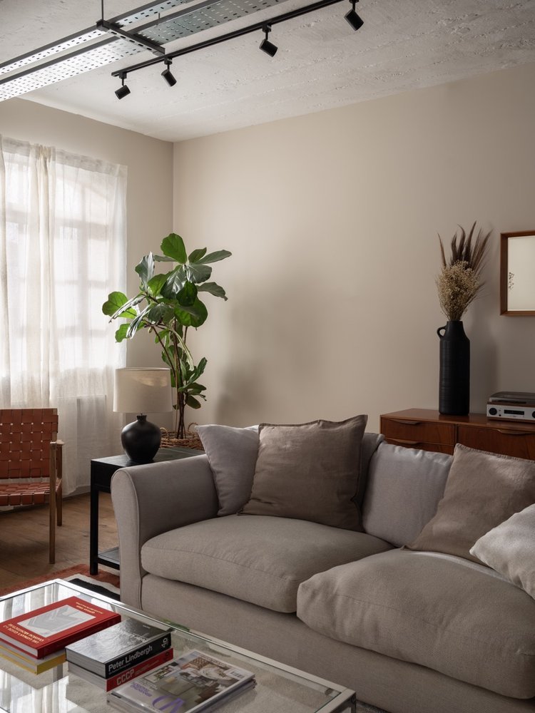 Living room with beige sofa, beige and gray cushions, glass coffee table, books, a black side table with a white lamp, large green potted plant, window with white curtains, wooden chair, black vase with dried plants, wall-mounted shelf with stereo eq