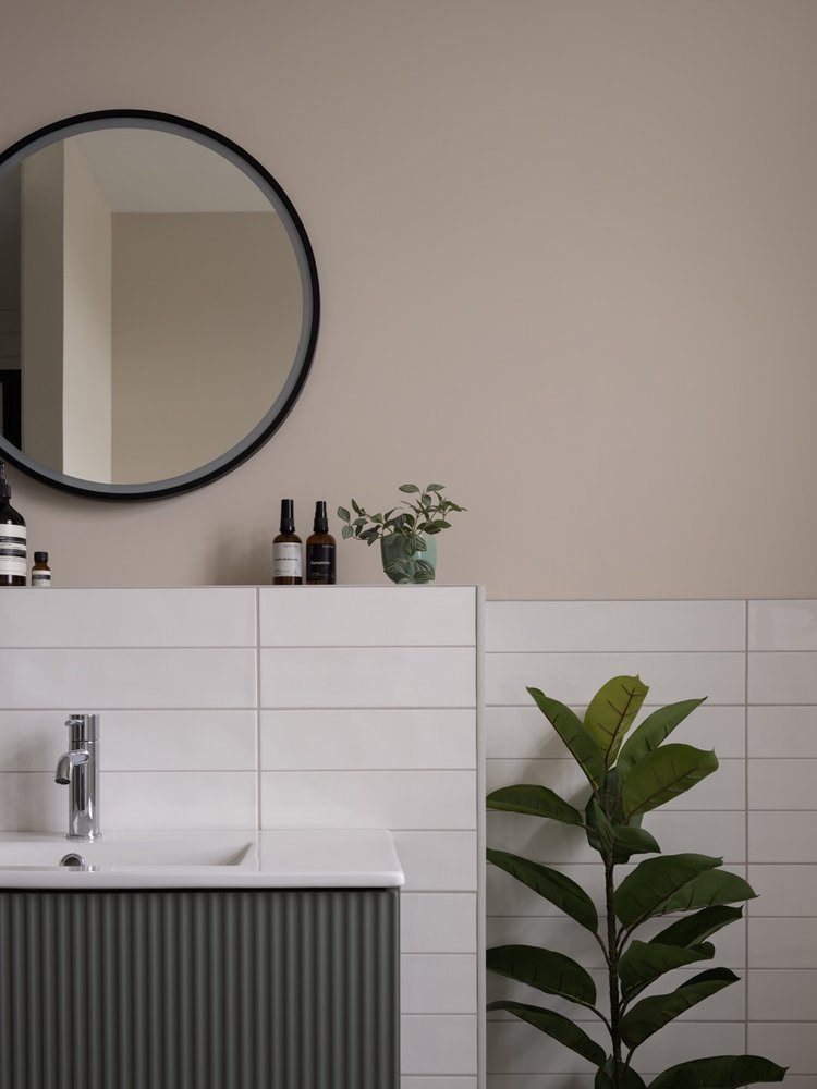 Bathroom with a black round mirror, white tiled wall, a small plant on top of the white tiled half-wall, a larger plant on the floor, and three amber bottles on the top of the wall.