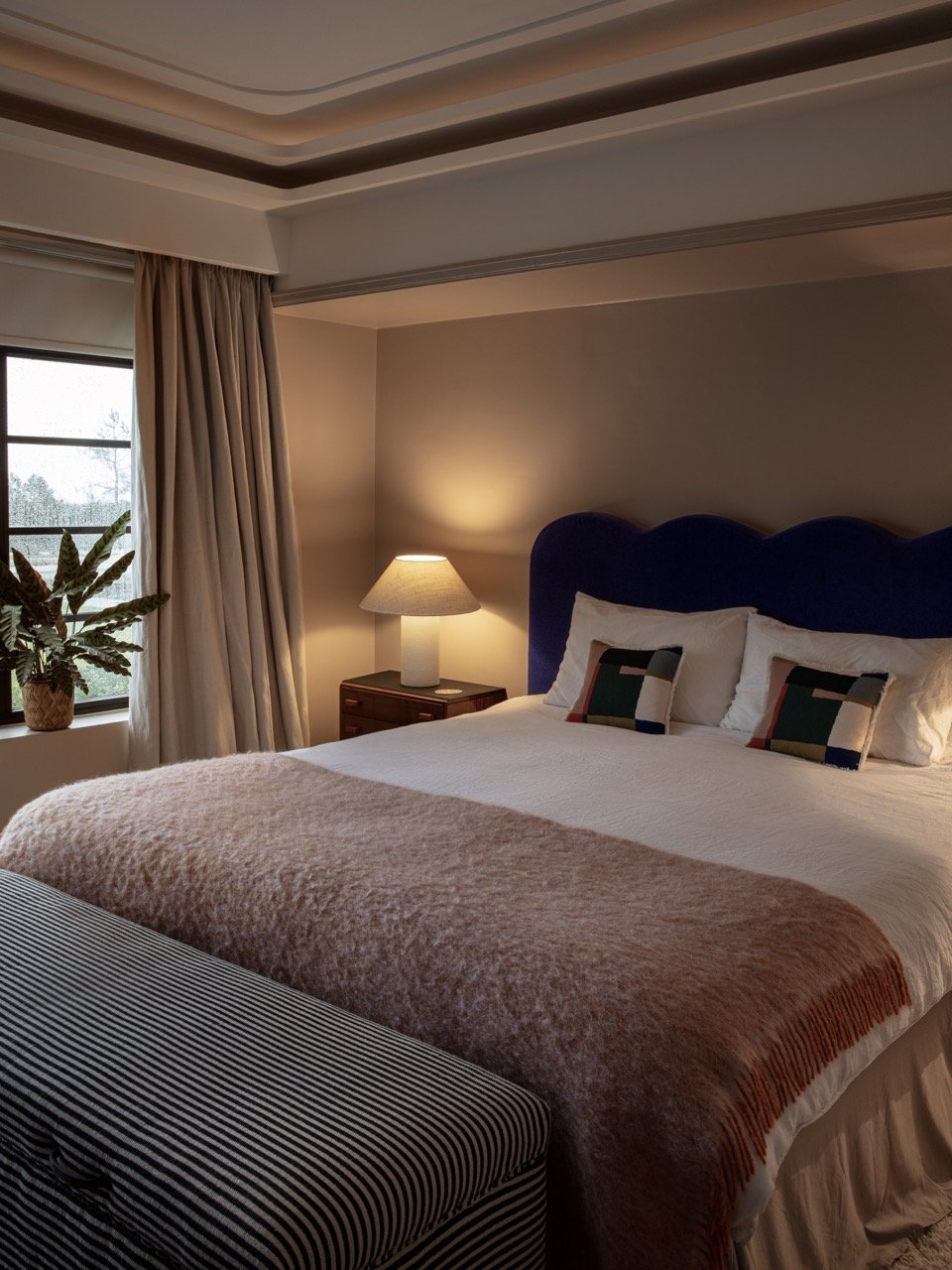 A cozy bedroom with a large bed that has a dark blue headboard, white bedding, and decorative pillows. There is a bedside table with a lamp, a window with curtains, and a potted plant in the corner.