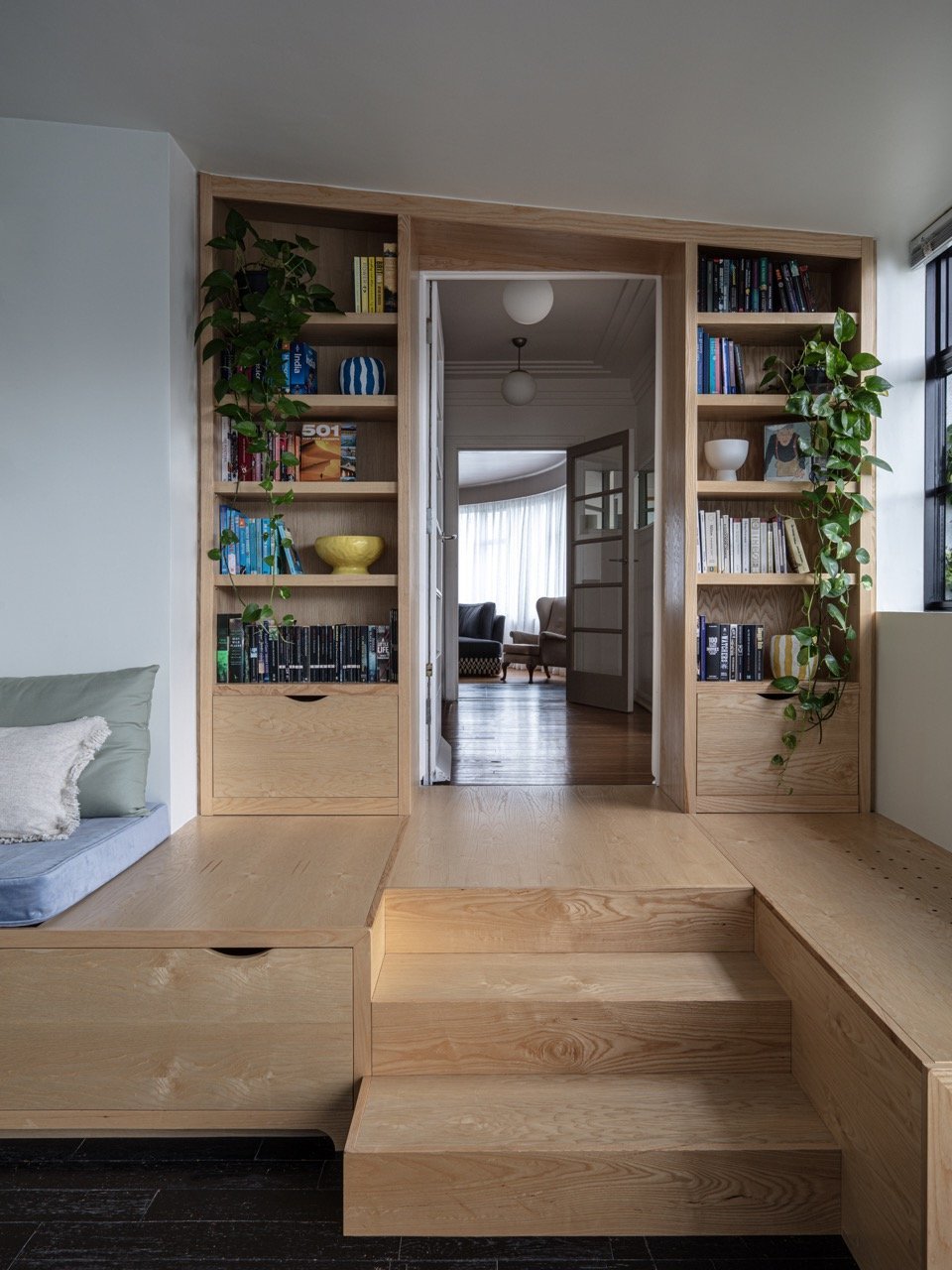 Wooden built-in shelf with books and plants, leading to a living room with large windows and curtains.