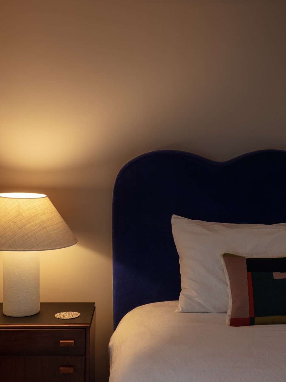 Close-up of a bedroom nightstand with a lamp and a decorative coaster, part of a bed with a dark blue upholstered headboard, white bedding, and a pillow with a colorful stripe pattern, against a beige wall.
