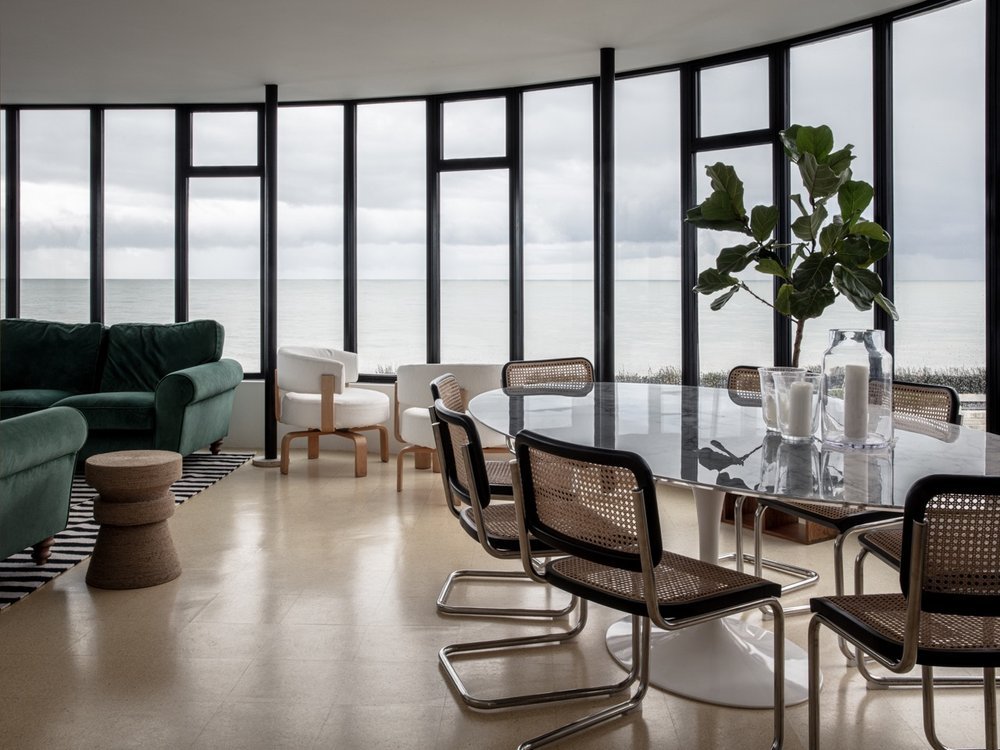 A modern living room with floor-to-ceiling windows overlooking the ocean, featuring a green sofa, white armchairs, a marble dining table with black chairs, and a large green plant.