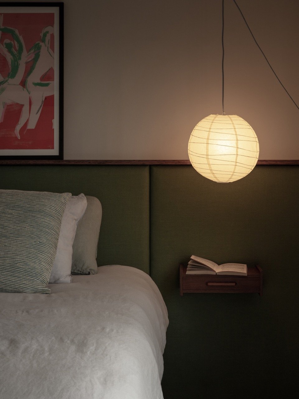 A cozy bedroom corner featuring a bed with white and gray striped pillows, a green upholstered headboard, a floating wooden bedside shelf with an open book, a hanging paper lantern, and a framed abstract artwork on the wall.