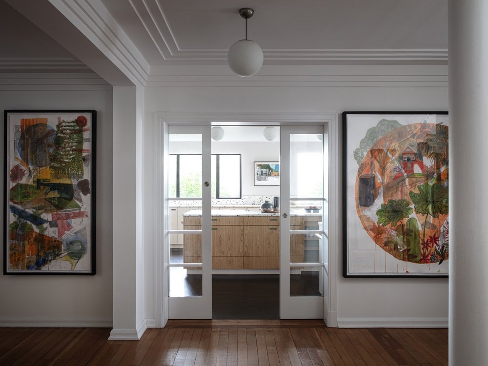 Interior view of a home with a doorway leading to a kitchen, featuring colorful abstract art paintings on either side of the door, wooden floors, and a ceiling light fixture.