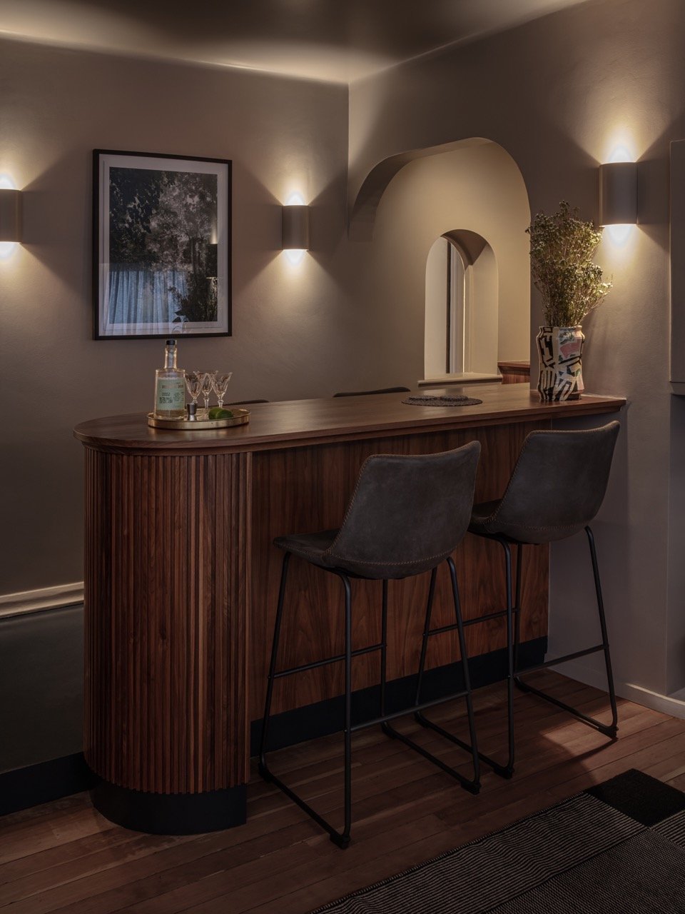 Dimly lit home bar with wooden countertop, two bar stools, and wall-mounted lights on beige walls. There is a framed picture on the wall, a bottle with glasses on a tray, and a vase with flowers on the counter.
