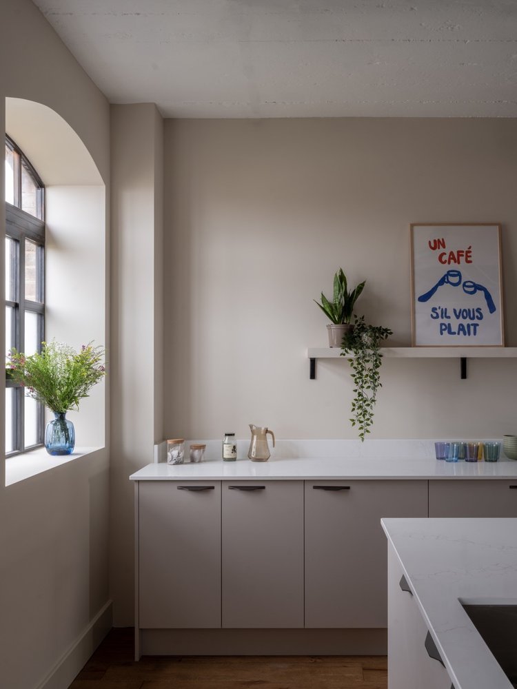 Minimalist kitchen with white cabinets, a window with flower vase, potted plants, and a framed sign on the wall that reads 'Un café, si vous plait.'