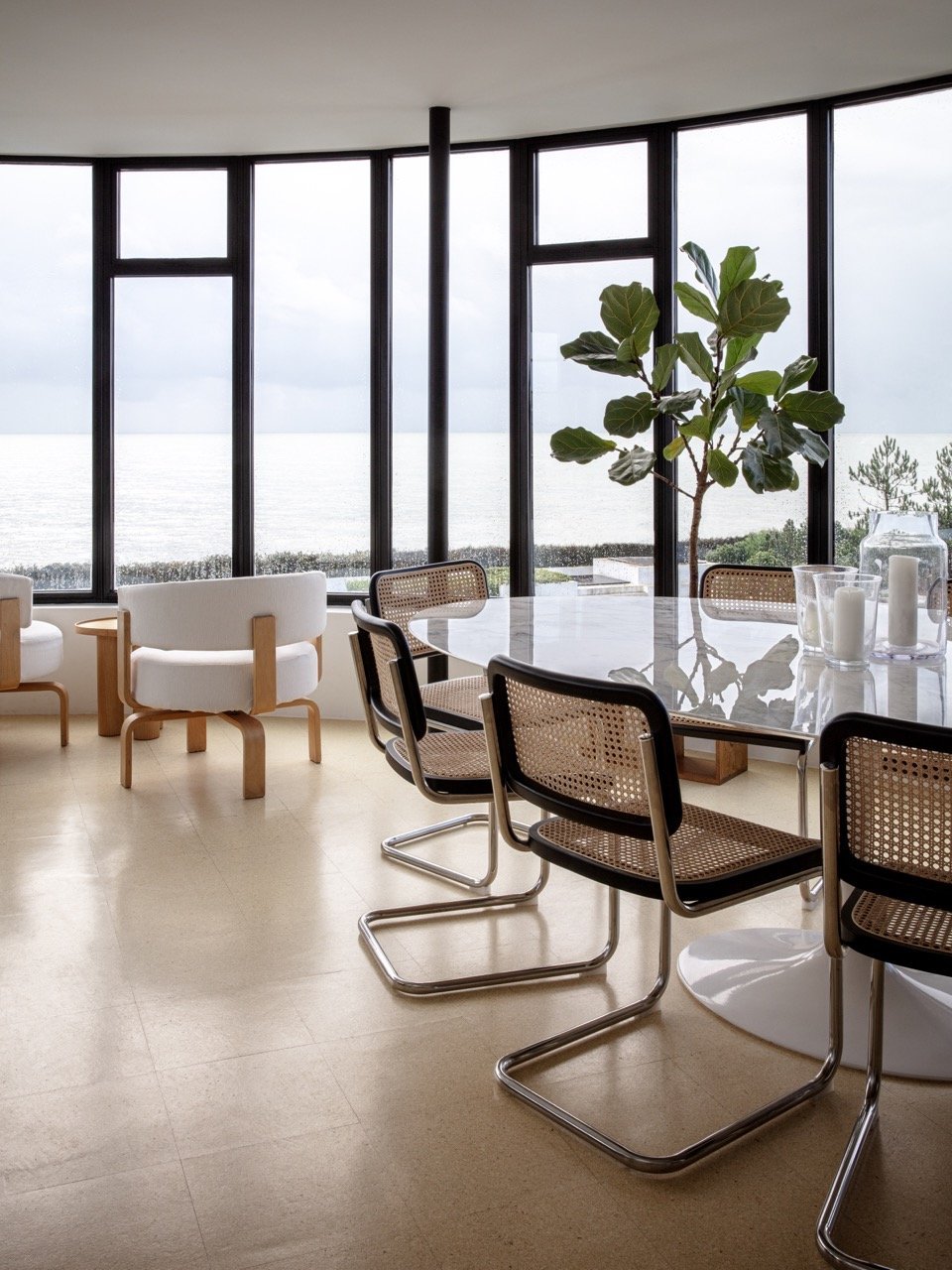 Interior of a modern room with large windows overlooking the ocean, a round glass table, several black and rattan chairs, a large indoor plant, and candles on the table.