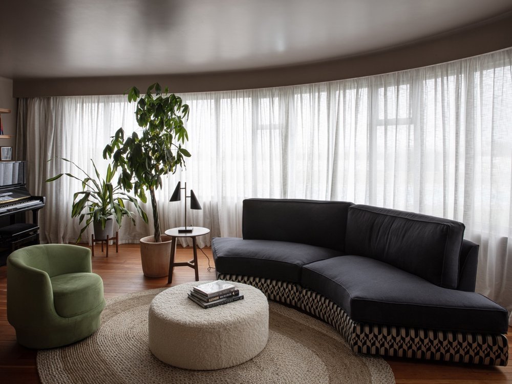 Living room with large curved window, sheer curtains, black sectional sofa, green armchair, round beige ottoman, potted plants, small side table with lamps, wooden flooring, and a black piano in the corner.