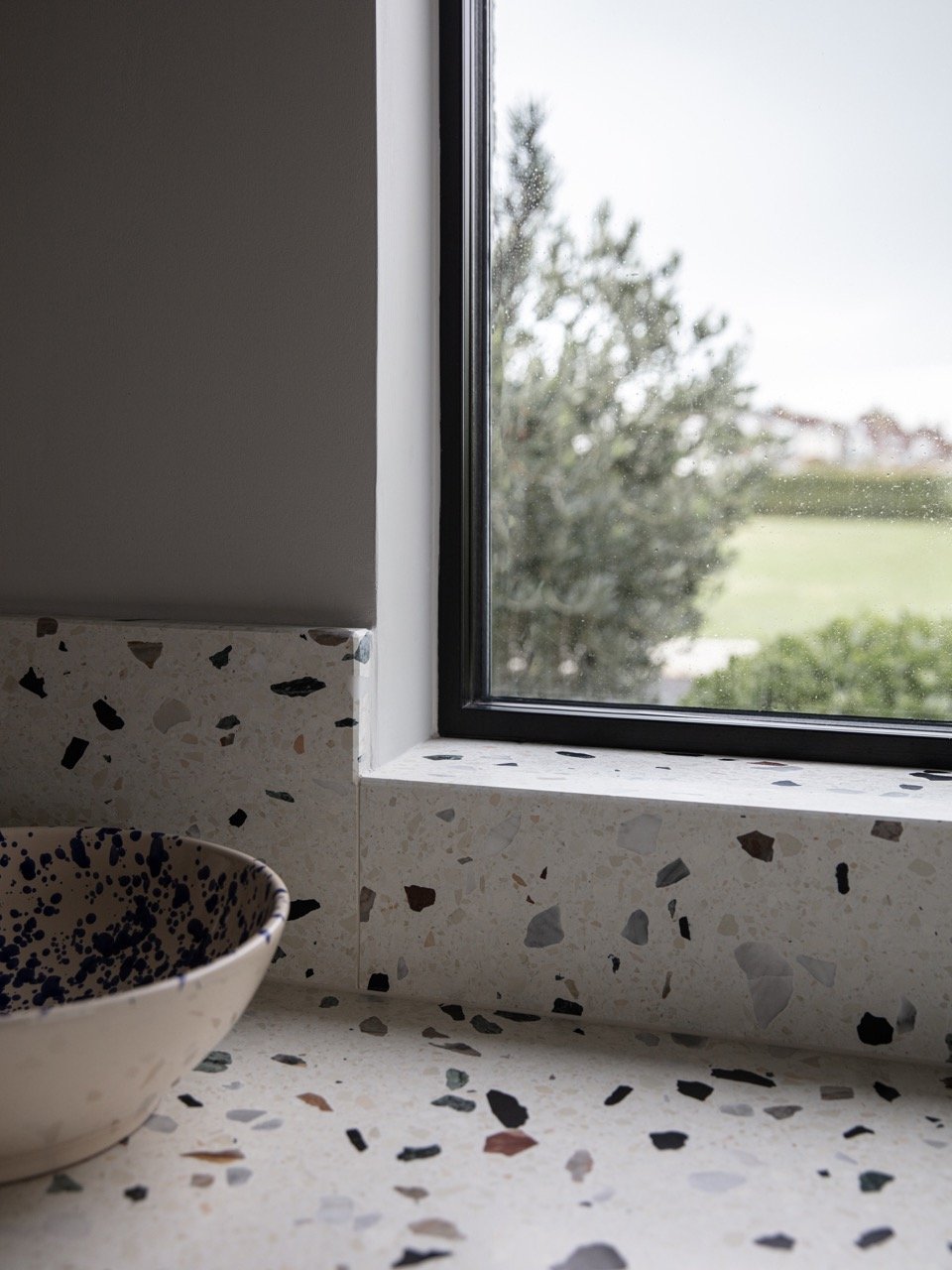 Close-up of a terrazzo countertop and windowsill with a ceramic bowl, overlooking a green outdoor landscape with trees and a cloudy sky.