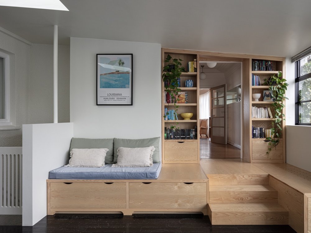 Interior of a modern living space with built-in wooden seating, a bookshelf, and potted plants, with a framed Louisianan poster on the white wall.