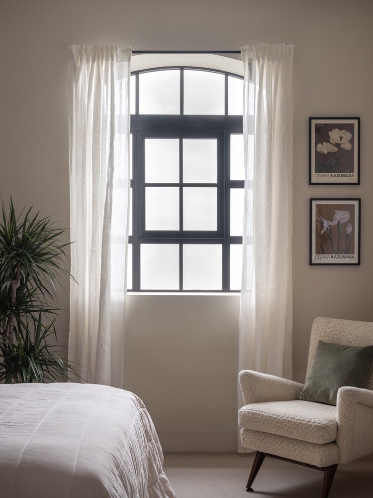A bedroom with a large arched window with black framing, sheer white curtains, a bed in the lower left corner, a beige armchair with a green pillow, and framed botanical prints on the wall.