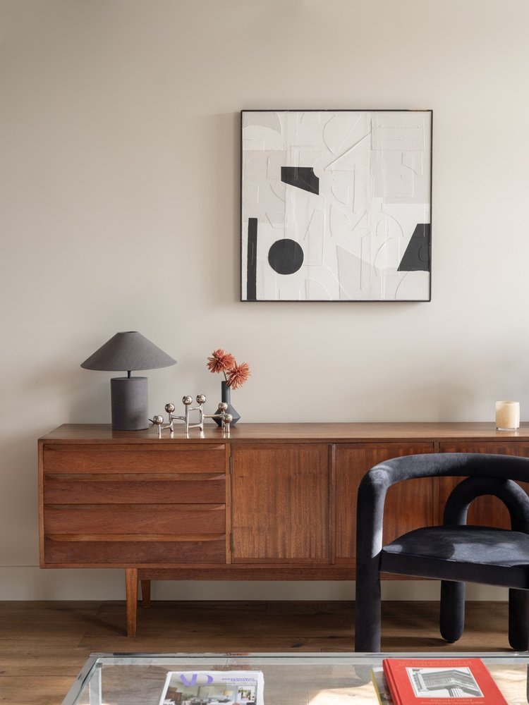 A mid-century modern wooden sideboard with decorative items including a gray lamp, a small vase with orange flowers, and metallic ball sculptures. Above it hangs a white abstract art piece with black geometric shapes. Part of a black upholstered chai