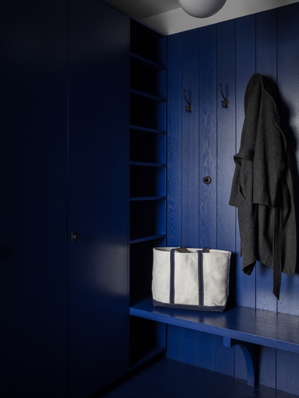 Blue wooden closet with open shelves, hooks with hanging coats, and a white tote bag with black straps on a blue bench.