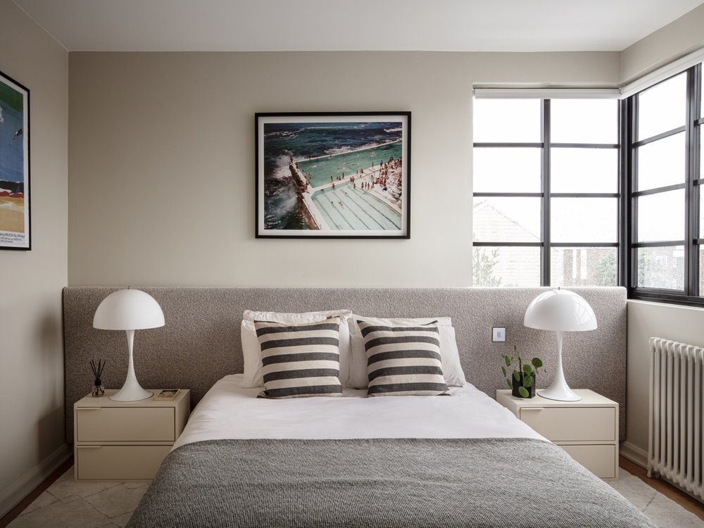 Bedroom with a bed, two nightstands with white lamps, striped pillows, framed artwork, and large windows.