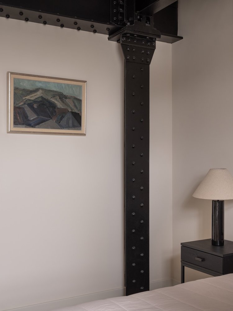 Interior of a bedroom with a large black metal support beam in the corner, a framed landscape painting on the wall, a nightstand with a lamp, and part of a bed with white bedding.