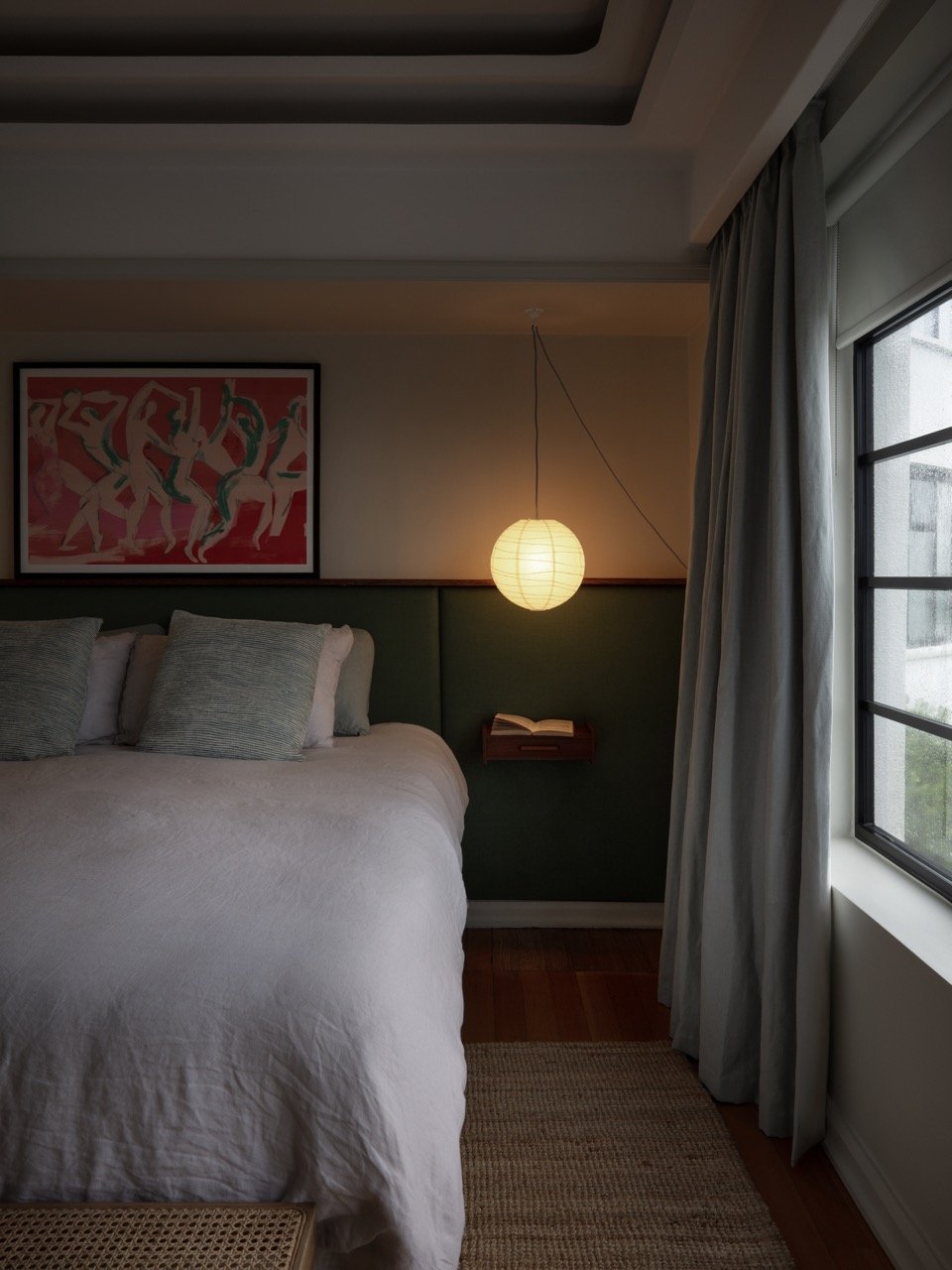 A cozy bedroom with a bed, pillows, a hanging paper lantern, a window with curtains, and a colorful artwork on the wall.