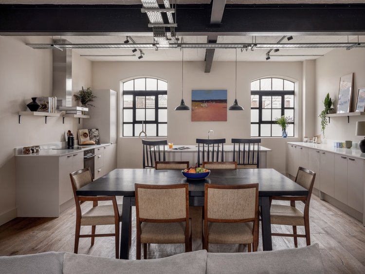 Modern dining room with a dark wooden table, six beige upholstered chairs, white kitchen cabinets, open shelves, two arched windows, and minimal decor including a centerpiece bowl of fruit and framed artwork.