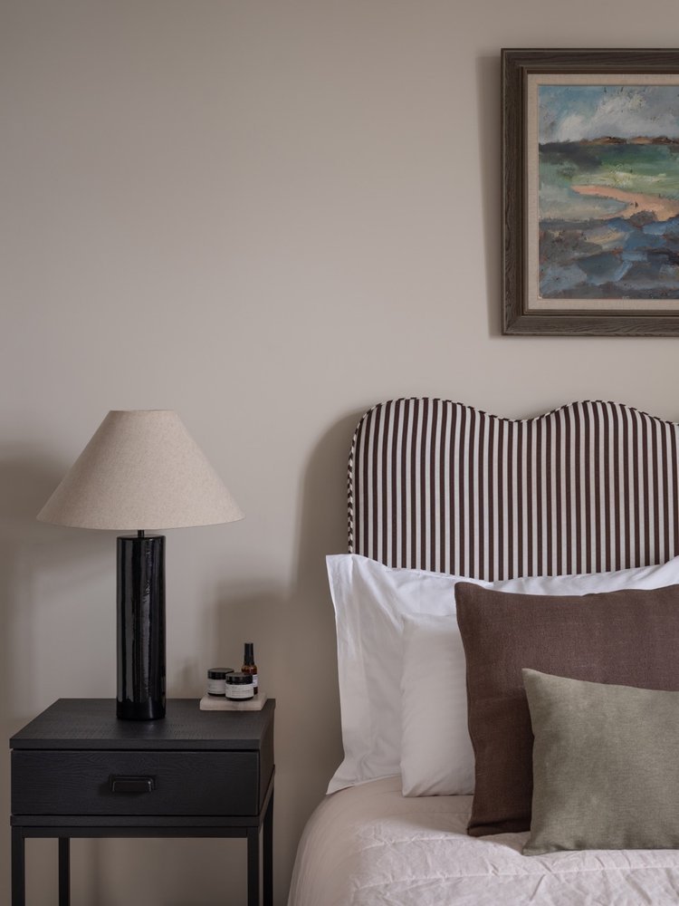 A bedroom with a striped upholstered headboard, pillows, a bedside table with a lamp, some small bottles, and a framed landscape painting on the wall.