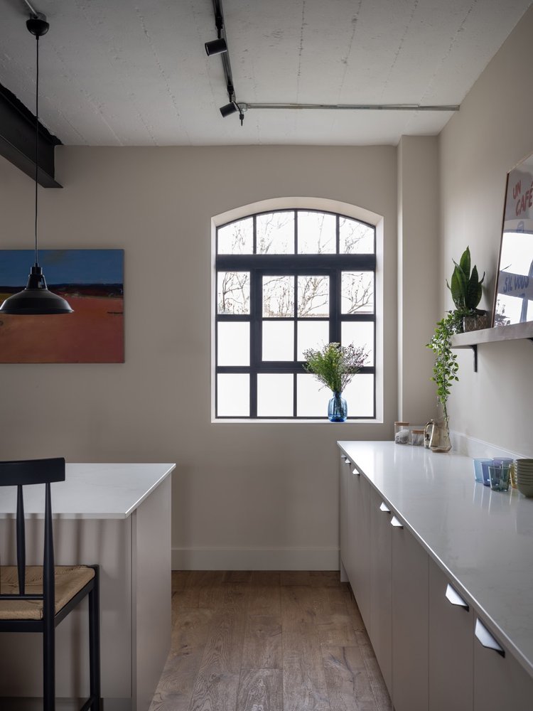 A minimalist kitchen with a large arched window, white cabinets, and a white countertop. A blue vase with flowers is on the window sill, and a small potted plant is on the wall shelf. A framed picture hangs on the wall, and a black pendant light is a