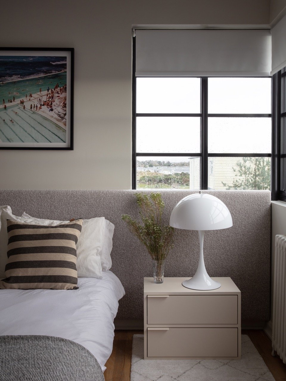A bedroom with a bed featuring a striped pillow, a nightstand with a glass vase of green foliage and a white modern lamp, a window with a white shade, and a framed picture of a beach scene on the wall.