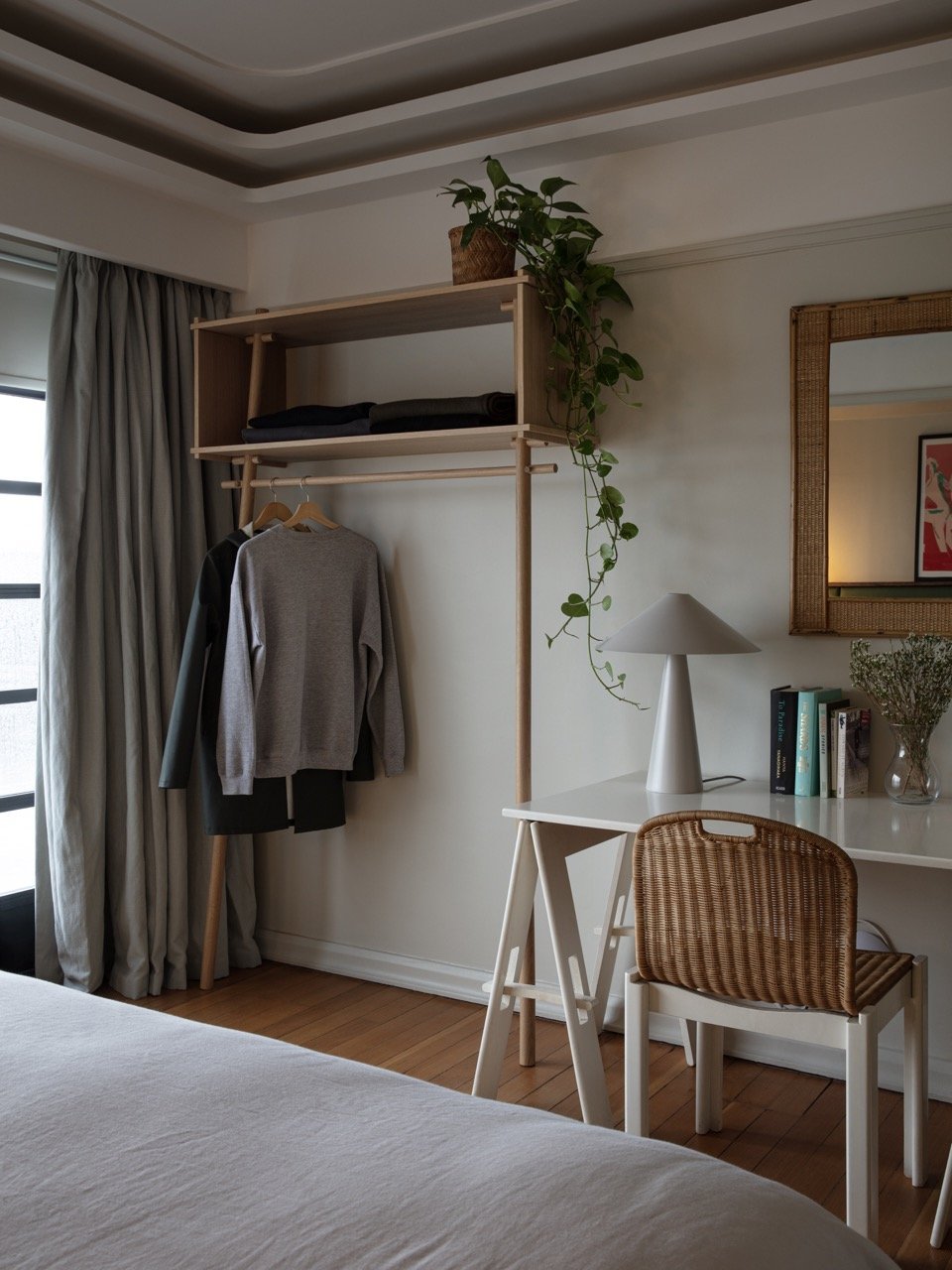 Bedroom corner with a white desk, wicker chair, white lamp, potted plant, books, mirror, and a clothes rack with folded towels and hanging garments, next to a window with curtains.
