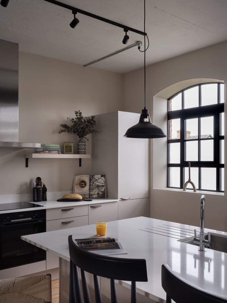 Modern kitchen with white cabinets, a black pendant light, a window with black framing, a white countertop, a glass of orange juice, and a black chair.