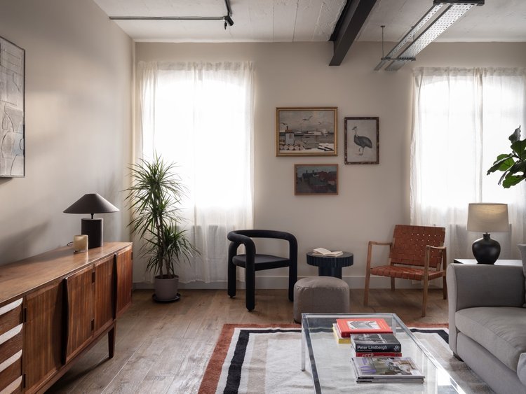 Living room with large windows, wooden sideboard, table lamp, potted plant, black and brown chairs, framed artwork, small side table, upholstered chair, sofa with a vase, and a glass coffee table with books.