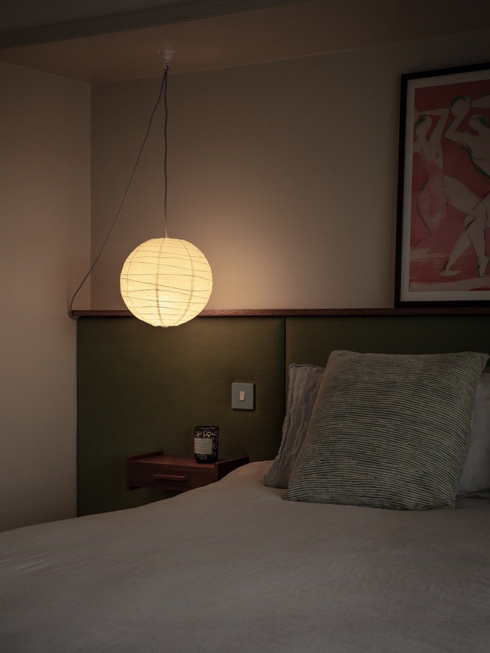 A cozy bedroom with a bedside lamp, a green upholstered headboard, a bedside table with a decorative item, and a framed artwork on the wall.