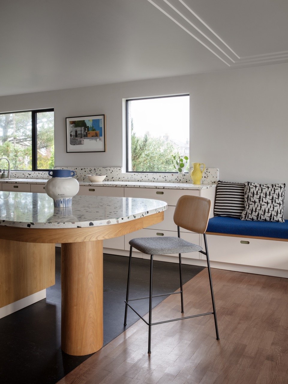 Modern kitchen with white walls, large windows, and a terrazzo countertop. There is a tall wooden stool with a gray cushion, a built-in bench with black and white pillows, and decorative vases on the countertop.
