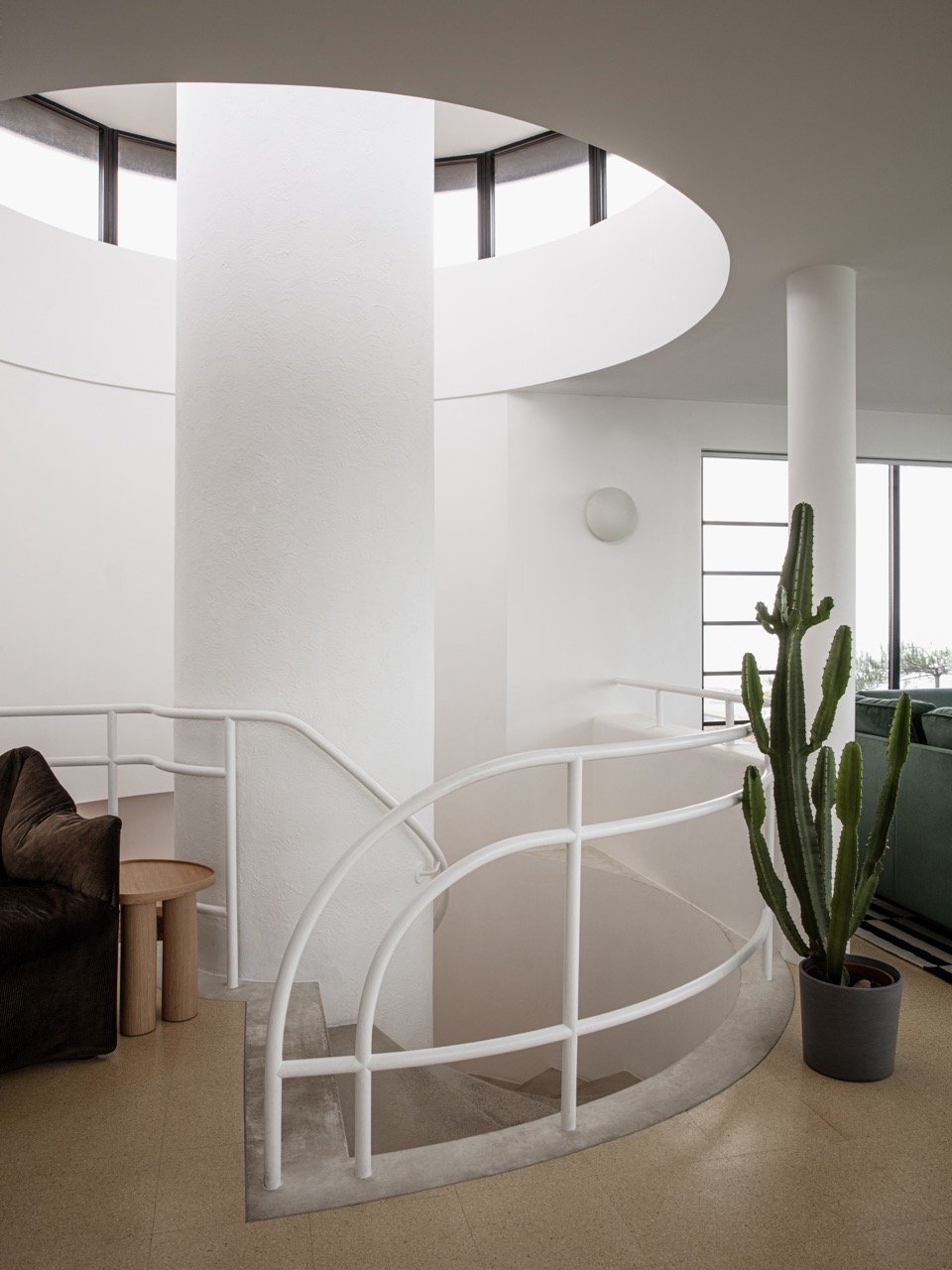 Interior of a modern home featuring a white spiral staircase, large windows, a tall cactus plant, a sofa, and a small wooden side table.