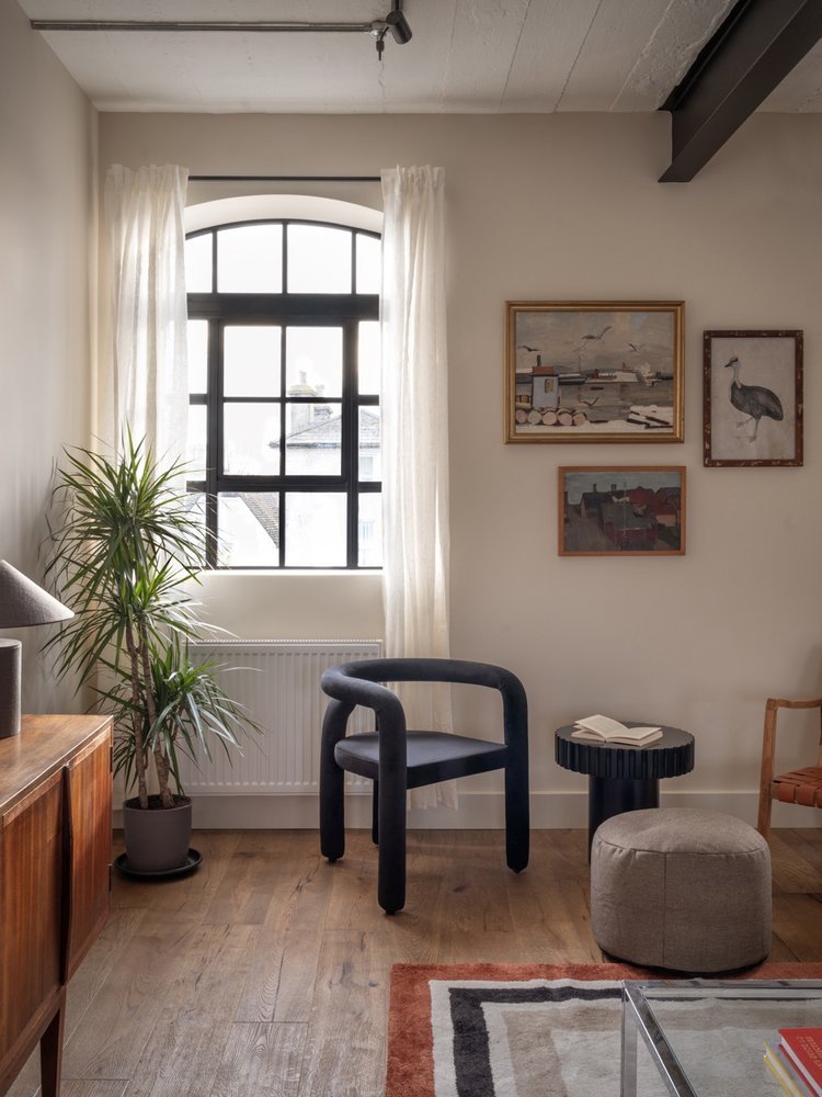 A cozy living room with a large arched window, white curtains, a potted plant, a black modern chair, a round ottoman with an open book, a small black side table, framed artwork on the wall, and part of a wooden cabinet and a patterned carpet.