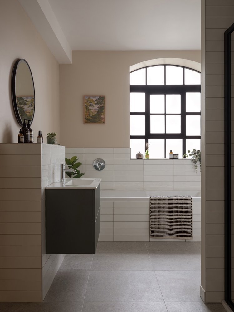 Bathroom with large arched window, white tile bathtub, black vanity with white sink, round mirror, plants, and small decorative items.