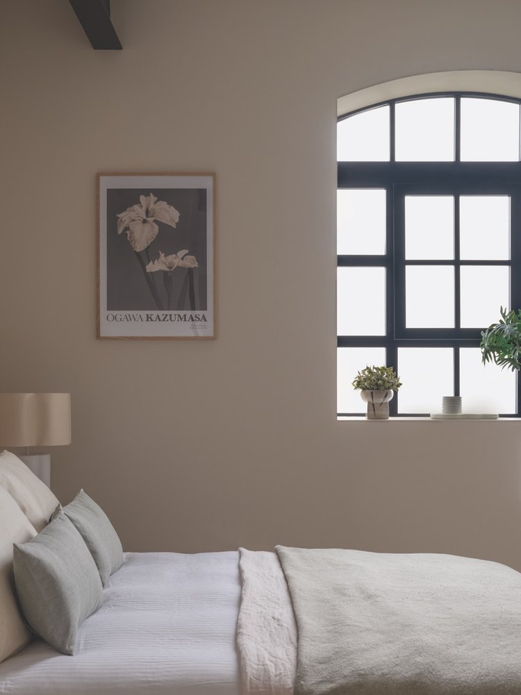 A bedroom with a bed, beige bed linens, two pillows, a beige wall, a framed flower art print, a black grid window with white panes, and a small potted plant on the window sill.