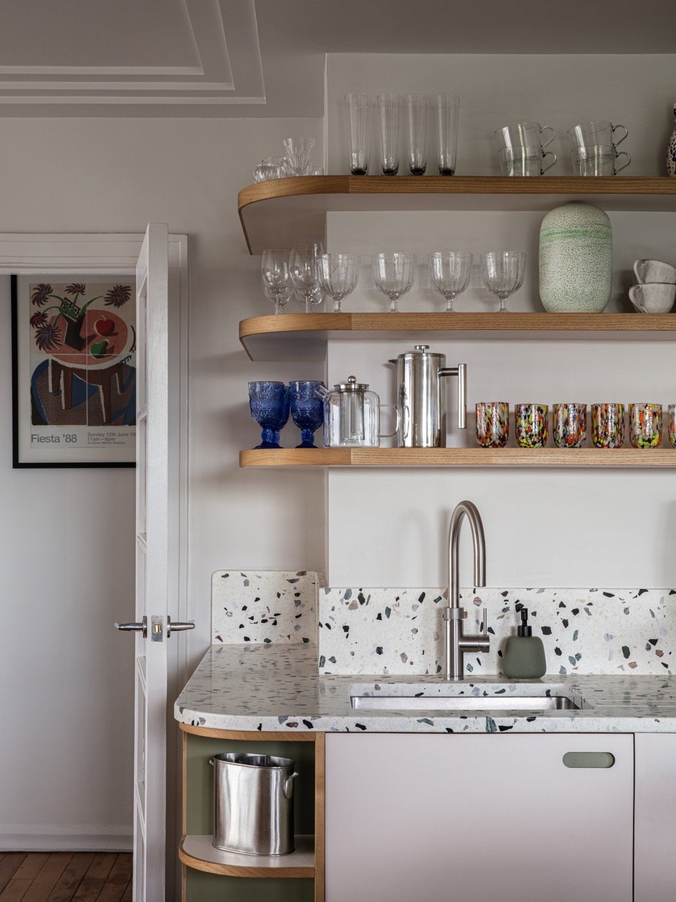 Kitchen with terrazzo countertop, open wooden shelves holding various glassware, a modern sink, and a small section of a door on the left side.
