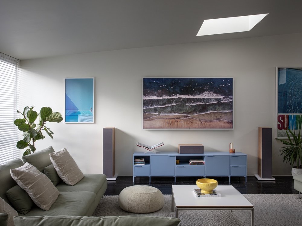 Modern living room with light green sofa, beige pillows, potted plant, large window with blinds, blue TV stand, large abstract ocean painting, smaller artworks, and a white coffee table with a yellow bowl.