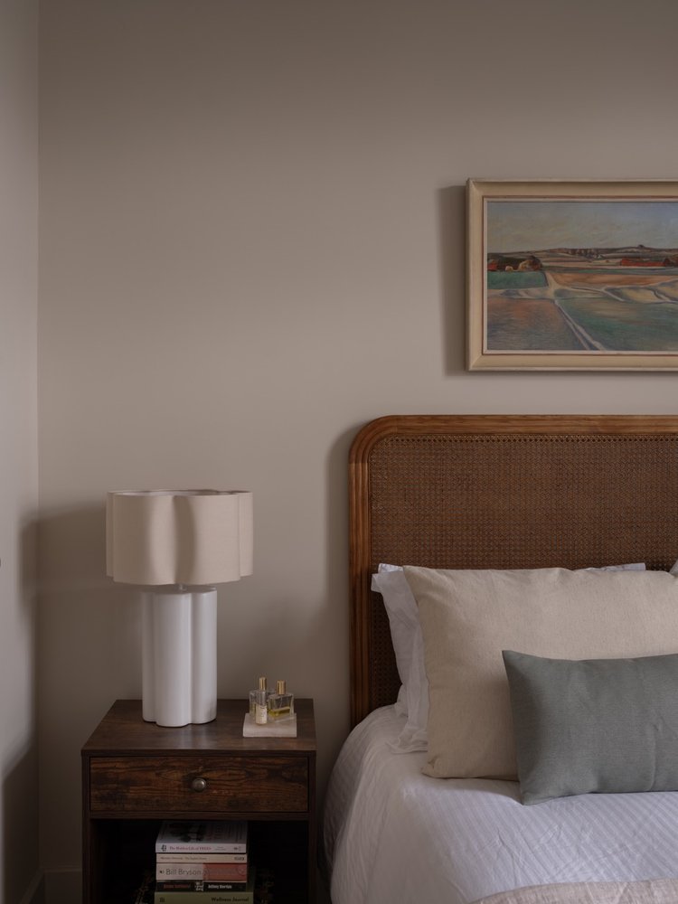 A bedroom corner with a wooden bed frame, white bedding, and a gray pillow. A wooden nightstand with a white lamp, perfume bottles, and books underneath. A framed landscape painting hangs above the bed.